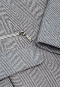 Un tissu en chevron gris avec une texture lisse présente une fermeture éclair argentée et un tirette métallique. Le motif est uniforme sur l'ensemble du matériau.