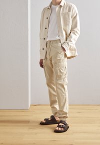 Giacca camicia in corduroy beige, t-shirt bianca, pantaloni cargo beige con tasche laterali e sandali con fibbia marroni. Sfondo in legno.