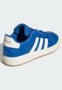 Modrá syntetická teniska s bílým designem tří pruhů, zaobleným tvarem, zavazováním vpředu a texturovanou gumovou podrážkou. Logo značky na patě.