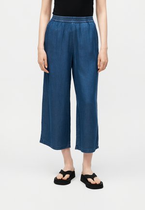 Persona che indossa pantaloni corti in denim blu a gamba larga con vita elastica e sandali neri con plateau su sfondo neutro.