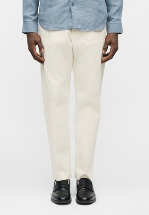 Homme portant un pantalon beige, des chaussures en cuir noires et une chemise bleue boutonnée, debout devant un fond clair uni.