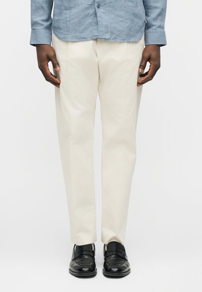 Homme portant un pantalon beige, des chaussures en cuir noires et une chemise bleue boutonnée, debout devant un fond clair uni.