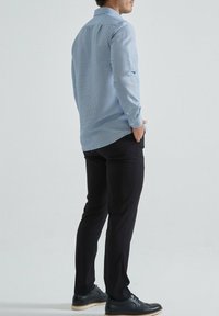 Chemise bleu clair à motifs, à manches longues et avec boutons, associée à un pantalon noir et des chaussures foncées, vue de dos.