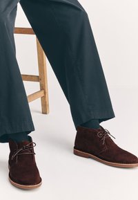 Braune Chukka-Stiefel aus Wildleder mit abgerundetem Zeh und braunen Schnürsenkeln, kombiniert mit dunklen Hosen und grauen Socken, neben einem Holzstuhl sitzend.
