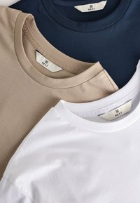 Trois t-shirts à col rond pliés en bleu marine, beige et blanc, chacun avec une étiquette visible de la marque NEXT à l'intérieur du col.