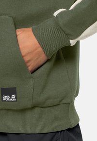 Ruka v kapse zelené mikiny Jack Wolfskin s ribbovaným manžetem a logem na přední kapse.