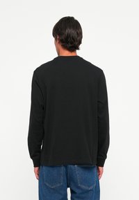 Mand med mørkt hår iført en ensfarvet sort langærmet skjorte og blå jeans, vendt væk mod en hvid baggrund.