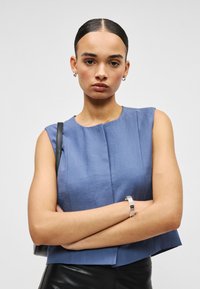 Γυναίκα με μαλλιά χτενισμένα προς τα πίσω, φοράει αμάνικο μπλε τοπ, μαύρο δερμάτινο παντελόνι, ασημένιο βραχιόλι και μικρούς κρίκους σκουλαρίκια, με τα χέρια σταυρωμένα.