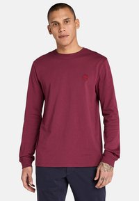 Langärmliges bordeauxrotes Baumwoll-T-Shirt mit Rundhalsausschnitt, ausgestattet mit einem kleinen roten Logo auf der Brust und einem gerippten Bündchen. Glatte Textur und gerade Passform.
