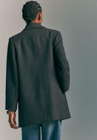 Persona con capelli intrecciati che indossa un cappotto grigio scuro, jeans blu e un braccialetto borchiato, mostrata di spalle su uno sfondo semplice.