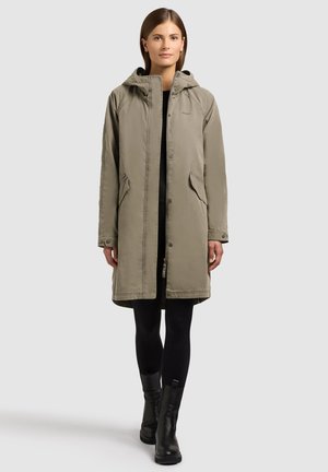 Žena nosící taupe parku po kolena s kapucí, černé legíny a černé kotníkové boty, stojící před světle šedým pozadím.