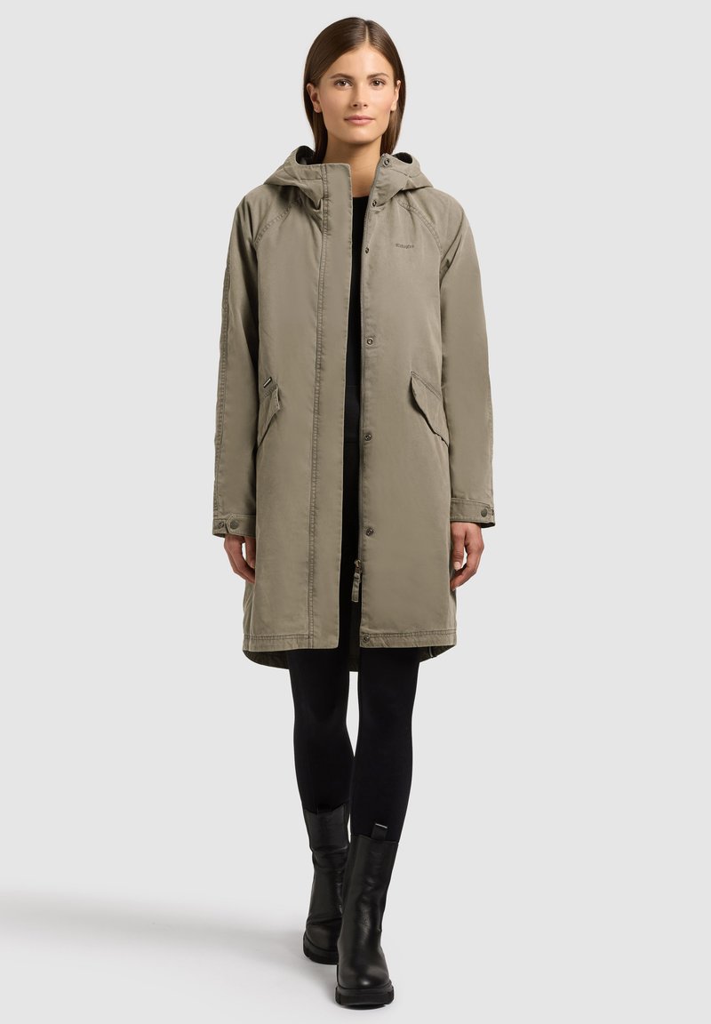 Femme portant une parka taupe longueur genou avec capuche, leggings noirs et bottines noires, debout devant un fond gris clair.