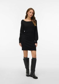 Žebrované černé šaty s dlouhým rukávem a čtvercovým výstřihem. Modelka je doplnila černými koženými kozačkami ke kolenům a šedými ponožkami. Hladká textura.