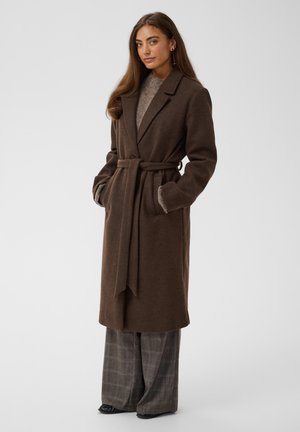 Femme aux longs cheveux bruns portant un manteau marron foncé ceinturé, un pull gris, un pantalon large à carreaux et des chaussures noires, debout les mains dans les poches.