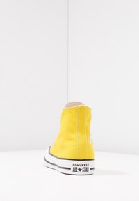 Gul højt snøresneaker af lærred med hvide sømme, sort gummisål og sorte detaljer. Har Converse All Star-mærkning på hælen.