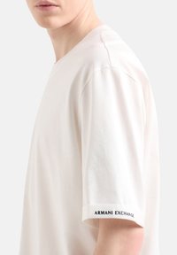 Rövid ujjú, krémszínű póló, puha pamutból készült, kerek nyakkivágással és a "ARMANI EXCHANGE" logóval fekete színben a hajtóka oldalán.