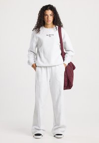 Mujer joven con cabello rizado que lleva un sudadera color gris claro de "New York City" y pantalones, llevando un bolso de hombro color burdeos, de pie sobre un fondo blanco.