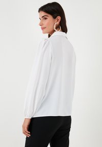 White long-sleeve blouse with a collared neck, gathered shoulders, and a smooth texture. Worn with black pants. Plain back view.