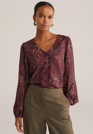 Femme portant un blouse à manches longues à motifs bordeaux, rentrée dans un pantalon tailleur taille haute vert olive, se tenant avec une main dans la poche.