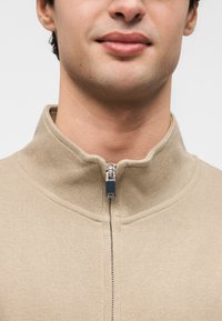 Μπεζ πουλόβερ με φερμουάρ και ψηλό γιακά, διαθέτει ασημένιο φερμουάρ και rib ύφασμα, δείχνοντας μια εφαρμοστή γραμμή γύρω από τον λαιμό.