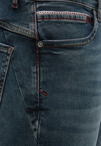 Nahaufnahme der Tasche einer dunkelblauen Jeans mit roter und gelber Naht sowie Details aus Metallnieten.