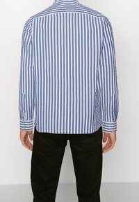 Sinivalkoraidallinen pitkähihainen paita, jossa on nappikaulus, kontrastiväriset kalvosimet ja rento istuvuus. Alhaalla näkyvät mustat housut.