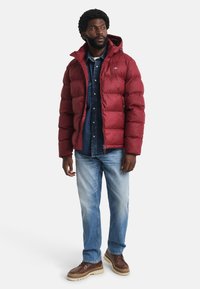 Mann in einer roten Daunenjacke über einem Jeanshemd und hellblauen Jeans, der steht und zur Seite schaut vor einem weißen Hintergrund.