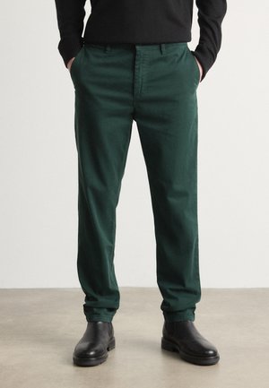 Personne portant un pantalon vert foncé droit, un haut noir à manches longues et des bottes en cuir noires, debout les mains dans les poches sur un sol uni.