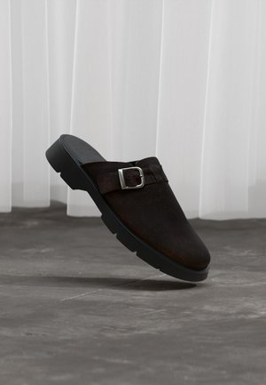 Mørkebrune slip-on træsko i ruskind med sort sål og sølvspænde, placeret i luften over en grå betongulv og hvidt gardinbaggrund.