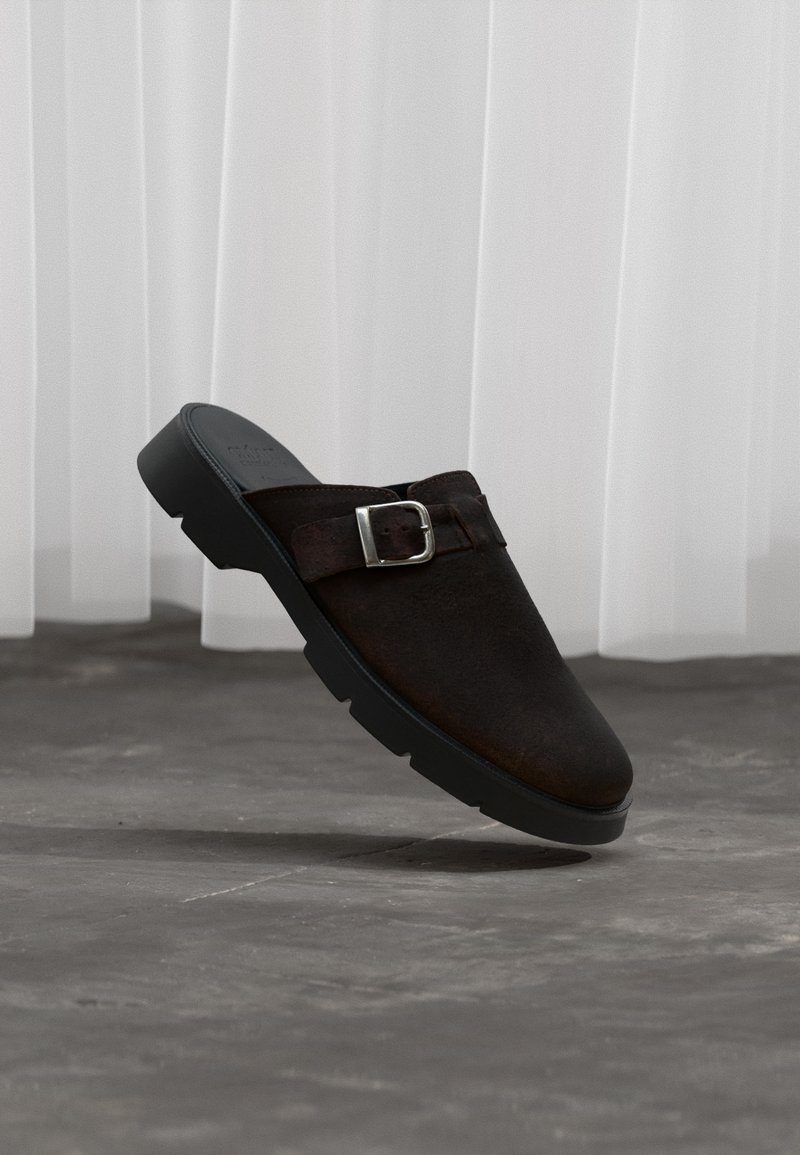 Σκούρο καφέ σουέτ παντόφλα χωρίς κορδόνια με μαύρη σόλα και ασημένιο λουράκι με πόρπη, τοποθετημένη στον αέρα πάνω από γκρι τσιμεντένιο δάπεδο και λευκό φόντο κουρτίνας.