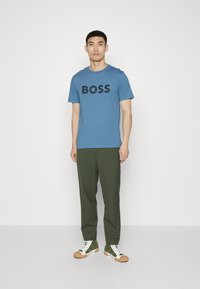 Kék, rövidujjú póló fekete "BOSS" felirattal, olajzöld nadrággal és zöld sportcipővel, fehér és barna részletekkel párosítva.