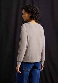 Lichtgrijze trui van gestructureerde stof, met ronde halslijn en geribbelde manchetten, gecombineerd met blauwe denim jeans. Model is van achteren afgebeeld.
