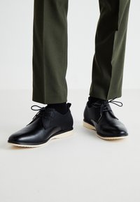 Scarpe in pelle nera con dettagli testurizzati, punta rotonda, suola beige a contrasto e chiusura con lacci, abbinate a pantaloni verde oliva.