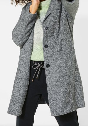 Osoba ubrana w szary teksturowany płaszcz na jasnozielonym swetrze z dzianiny i czarne spodnie z białym sznurkiem związanym w kokardkę.