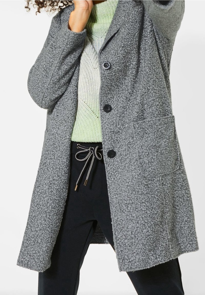 Personne portant un manteau gris texturé par-dessus un pull en tricot vert clair et un pantalon noir avec un cordon blanc noué en nœud.