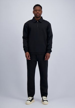 Man standing wearing a black long-sleeve quarter-zip top, matching black pants, and black and white sneakers on a plain background.