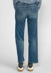 Jean en denim bleu foncé avec une coupe droite, dotés de deux poches arrière avec un motif de couture unique. Portés avec des baskets blanches.