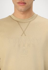Béžové tričko s okrúhlym výstrihom vyrobené z mäkkej bavlny, s embosovaným logom "TOMMY JEANS" a emblémom koruny s dekoratívnym prešívaním.