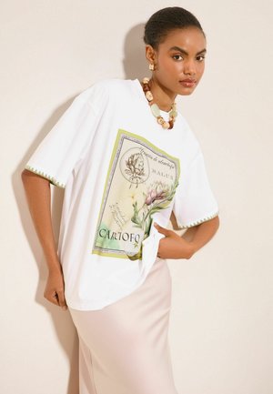 Kvinde iført hvid oversized t-shirt med botanisk kunst og beige nederdel, accessorized med chunky perlehalskæde og øreringe mod ensfarvet baggrund.