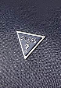 Matériau texturé bleu marine avec un logo triangulaire argenté portant "GUESS USA" et un point d'interrogation, sur un fond à motifs.