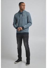 Sudadera azul claro con un bolsillo canguro en el frente y puños oscuros. Combinada con jeans oscuros y zapatos negros. Diseño simple y casual.