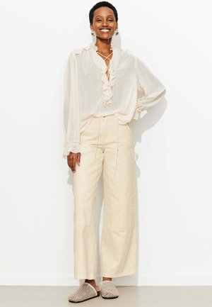 Femme souriante debout, main sur la hanche, portant un chemisier crème à volants, un pantalon pattes d'éléphant et des chaussures à enfiler tissées, sur fond blanc.