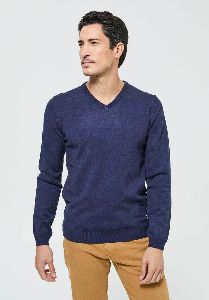 Pull-over à col en V bleu marine avec des manches longues, texture tricotée et poignets côtelés. Associé à un pantalon kaki. Design minimaliste, sans motifs.