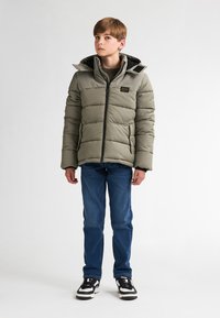 Giacca puffer khaki con cuciture orizzontali, cappuccio e tasche frontali. Abbinata a jeans blu e sneaker nere con dettagli bianchi.