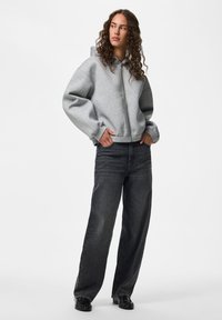 Donna con capelli ricci che indossa una felpa grigia con zip, jeans neri a gamba larga e scarpe nere, in piedi con le mani in tasca.