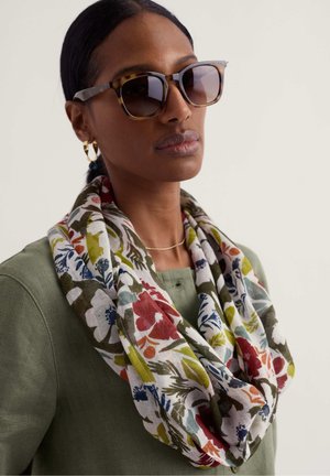 Femme portant des lunettes de soleil écaille, une boucle d’oreille créole en or, un collier fin, un haut vert olive et une écharpe à motif floral aux teintes rouges, bleues et vertes.