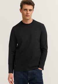 Mann trägt ein schwarzes Langarmshirt mit kleinem "Bugatti"-Logo auf der Brust und blaue Jeans, steht mit einer Hand in der Tasche.