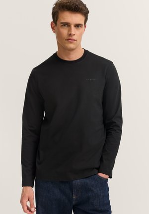 Mann trägt ein schwarzes Langarmshirt mit kleinem "Bugatti"-Logo auf der Brust und blaue Jeans, steht mit einer Hand in der Tasche.