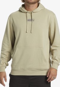 Sudadera beige hecha de tela suave, con un bolsillo frontal, cordones en la capucha y un pequeño parche con el logo centrado en el pecho.