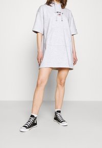 Šedé oversize šaty z mikiny s přední kapsou a logem. Nosí se s černými vysokými teniskami a bílými kotníkovými ponožkami. Hladká textura.